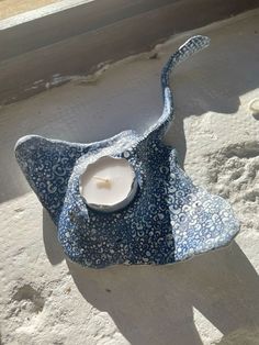 a blue and white flowered hat with a candle in it on the sand near a window sill