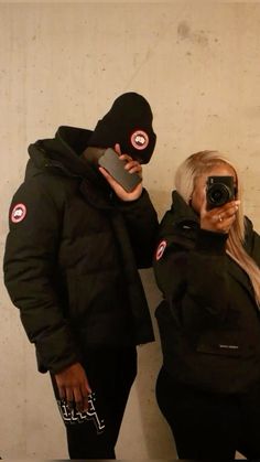 a woman taking a selfie in front of a man wearing a black jacket and hat