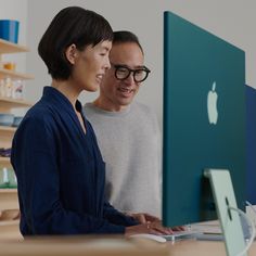 a man and woman looking at an apple computer on a desk in front of shelves