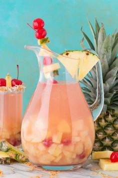 a pitcher filled with pineapple punch and garnished with cherries next to sliced pineapples