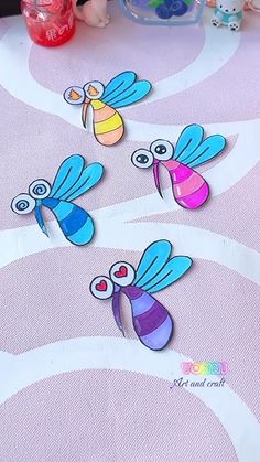 paper cut out of three dragonflies sitting on top of a pink tablecloth with other items in the background