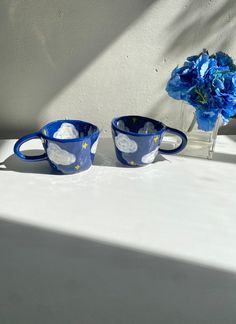 two blue cups sitting next to each other on a white table with flowers in it