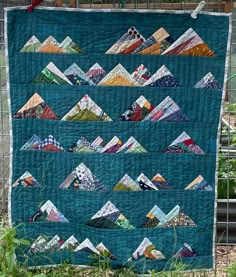 a quilt hanging from a wire fence in front of some plants and trees with mountains on it