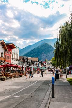 people are walking down the street in front of some buildings and trees with mountains in the background