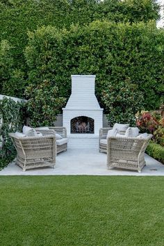 an outdoor fireplace surrounded by wicker chairs and shrubbery in a backyard area with grass