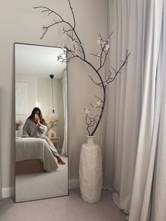 a woman sitting on a bed in front of a mirror with a branch and flowers