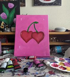 two cherries on a pink canvas surrounded by paint and crayons, with tulips in the background