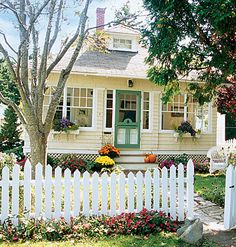 a white picket fence in front of a yellow house with flowers and trees around it