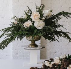 white flowers and greenery in a gold vase on a pedestal next to pine cones