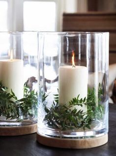 two clear glass vases with candles and greenery in them sitting on a table