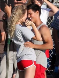 a man and woman hugging each other in front of a fence with people behind them