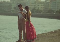 a man and woman standing next to the water looking at something on their cell phone