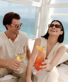 a man and woman sitting on a couch holding wine glasses with the ocean in the background