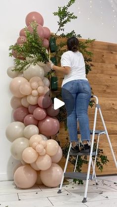 a woman standing on a ladder next to a wall with balloons and greenery in it