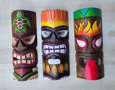 three tiki masks are lined up on a white surface, each with different designs and colors