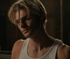 a man with blonde hair wearing a tank top in a dark room looking at the camera