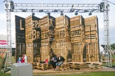 two people sitting on wooden pallets in front of an outdoor stage set made out of wood planks