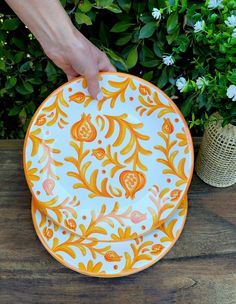 a hand holding an orange and white plate on top of a wooden table next to flowers