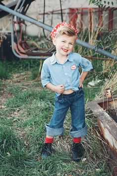 a young boy standing in the grass wearing jeans and a red bandanna on his head