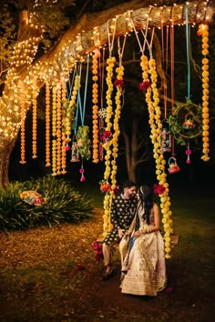 a man and woman sitting on a bench under a tree covered in yellow string lights