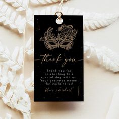 a black and gold thank you card hanging from a necklace on a white background with leaves