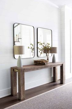 two mirrors on the wall above a wooden table with vases and lamp next to it