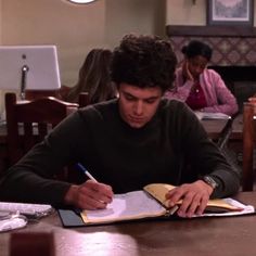 a young man sitting at a table writing in a notebook with two other people behind him