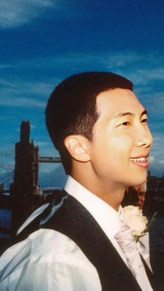 a man in a suit and bow tie standing next to the water with a bridge in the background