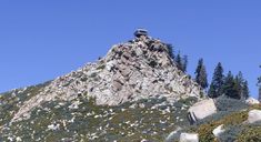 the top of a mountain with trees and rocks on it's sides, against a blue sky