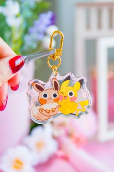 a hand holding a keychain with two cartoon characters on it and a flower pot in the background