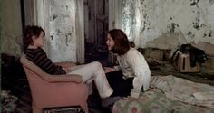 two women sitting on chairs in an old room with peeling paint and crumbling walls, one is talking to the other