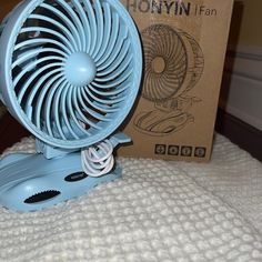 a small blue fan sitting on top of a bed next to a box with the lid open