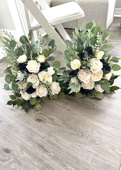 two white roses and greenery are sitting on the floor next to each other in front of a chair