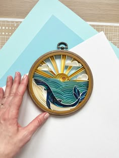 a hand is holding an embroidery project on top of some papers and paper with the image of a whale swimming in the ocean