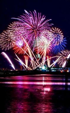 fireworks are lit up in the night sky over water and buildings with lights on them