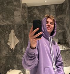 a man in a purple hoodie taking a selfie with his cell phone while standing in front of a bathroom sink