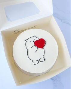 a white cake with a red heart on it sitting in a box next to a card