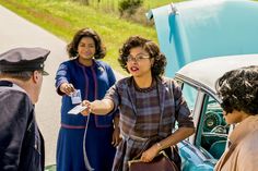 two women are talking to three men in front of an old car on the road