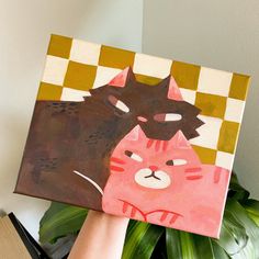 a person holding up a painting with a cat on it's face next to a potted plant