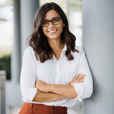 a woman wearing glasses leaning against a pillar with her arms crossed and looking at the camera