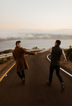 two people holding hands while walking down the road by the ocean in the evening time