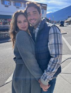 a man and woman are standing together in the middle of an empty street with mountains in the background