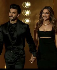 a man and woman holding hands while walking down a hall with lights on the wall behind them