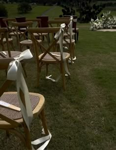 rows of wooden chairs with white ribbons tied to them on the grass in front of an open field