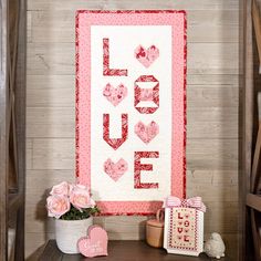 a valentine's day quilt hanging on a wall next to flowers and a potted plant