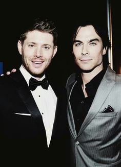 two men in tuxedos posing for a photo together at an event, one is wearing a suit and the other has a bow tie