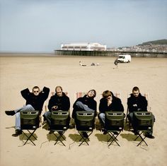 four people sitting in lawn chairs on the beach with their backs turned to the camera