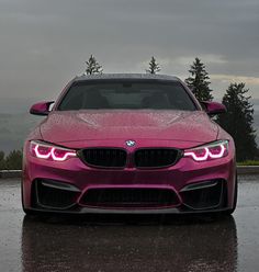 the front end of a pink bmw car on a wet road with trees in the background