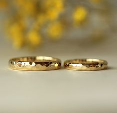 two gold wedding rings sitting next to each other on a white surface with yellow flowers in the background