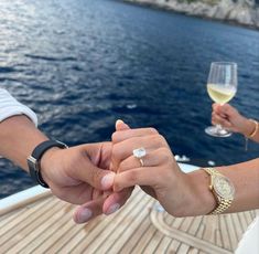 two people holding hands while standing on a boat in the water with wine glasses nearby